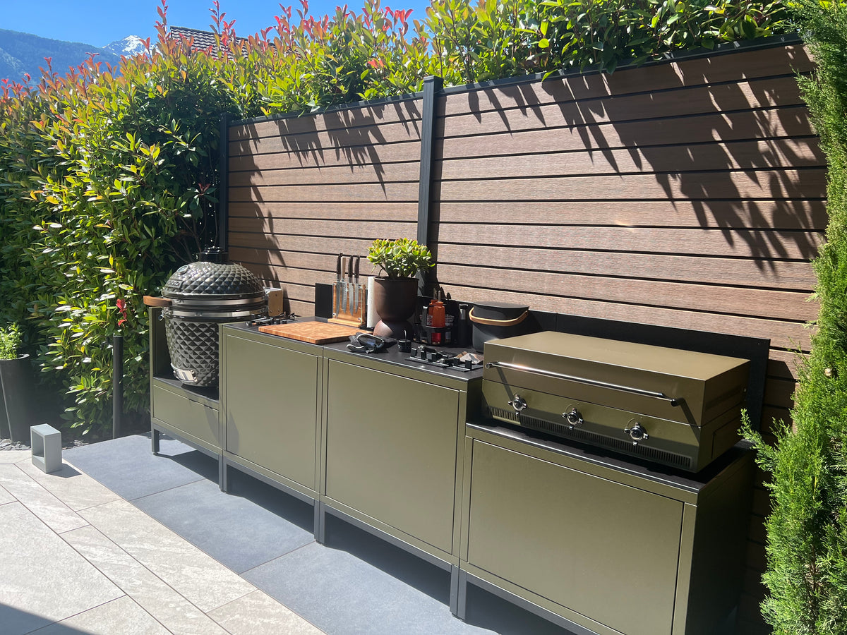 Vue de côté d'une cuisine extérieure modulaire kaki avec plancha au premier plan et barbecue kamado Kokko au fond, sur une terrasse ensoleillée avec vue sur les montagnes.